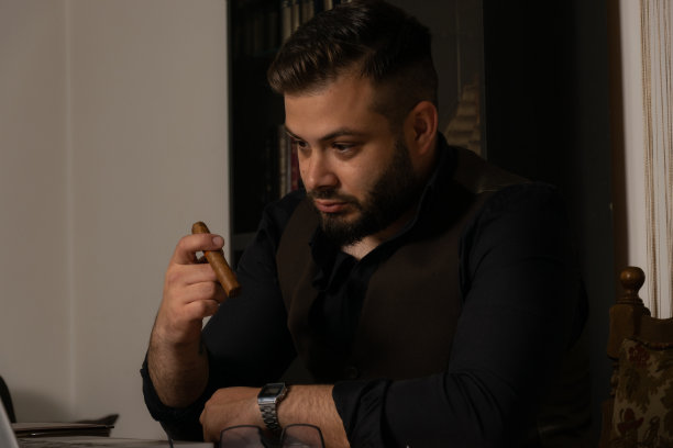 一个年轻的商人在工作了一整天后，抽着雪茄试图放松，同时检查他的工作在他的办公桌上的笔记本电脑。图片下载