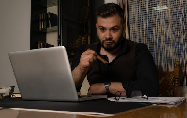 一个年轻的商人在工作了一整天后，抽着雪茄试图放松，同时检查他的工作在他的办公桌上的笔记本电脑。图片下载