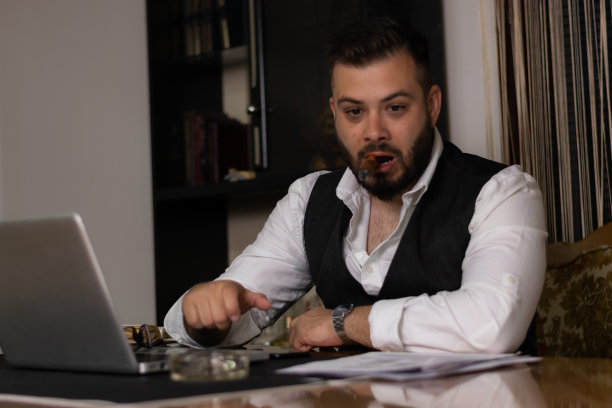 一个年轻的商人在工作了一整天后，抽着雪茄试图放松，同时检查他的工作在他的办公桌上的笔记本电脑。图片下载