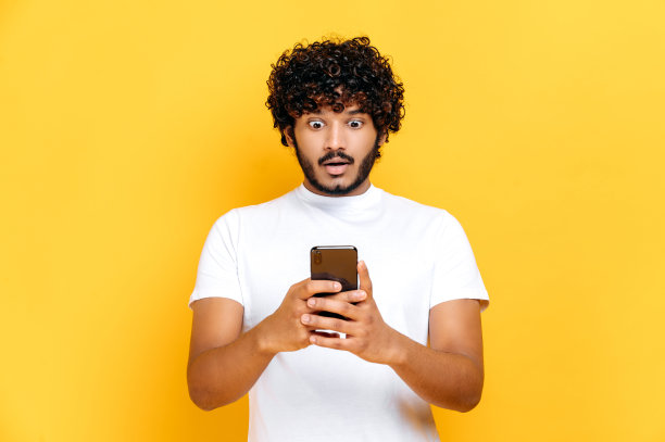 穿着白色t恤的印度人惊讶地用他的智能手机，惊讶地看着屏幕，收到了意外的信息或新闻，站在孤立的橙色背景上，表情困惑图片下载