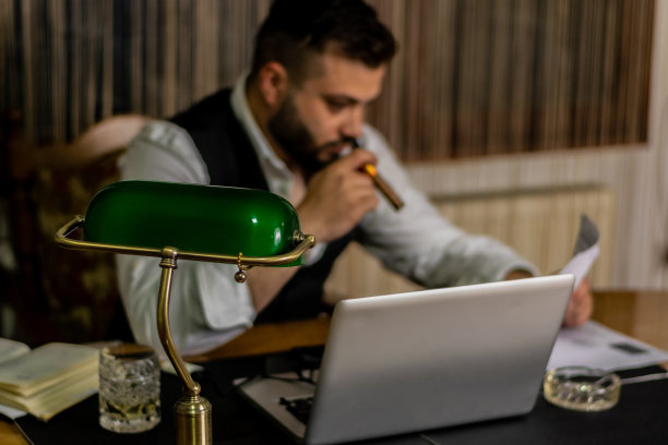 商人在他的办公室工作在他的办公桌上的笔记本电脑，在漫长的一天的办公室后，结束他的工作，抽着雪茄。图片下载