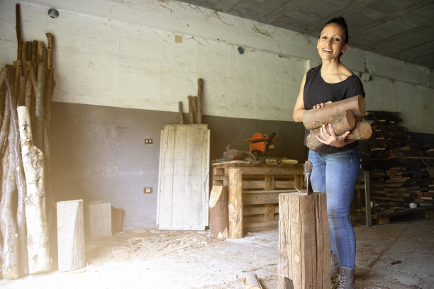 一个女人用木头砍斧头当柴火的水平照片。仓库里堆满了木头。在工作中微笑的女人被认为是男人的工作。图片下载