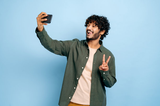 快乐积极的印度或阿拉伯人兴奋的家伙，穿着休闲时尚的衣服，用手机自拍，对着相机微笑，在孤立的蓝色背景上摆出和平符号的姿势图片下载