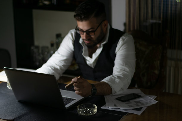 严肃英俊的商人，西装革履，倚在桌子上看图表。深夜工作的概念。工作到深夜为第二天做准备，完成他的工作。图片下载