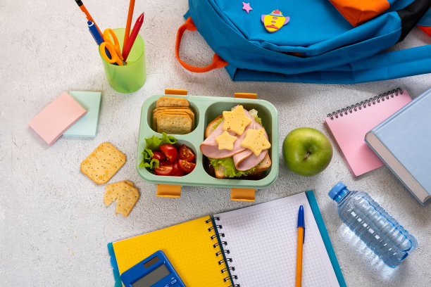 带三明治和沙拉的健康学校午餐盒放在工作地点背包附近。学校用品，书，苹果和一瓶水。图片下载