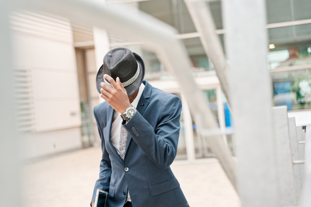 西装男把脸藏在帽子后面。商人是反映在办公楼的玻璃。图片下载