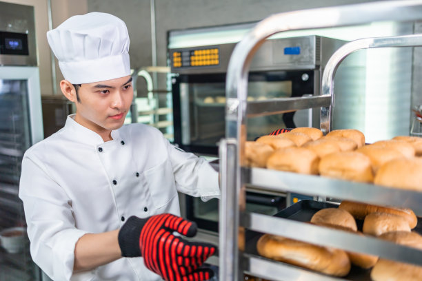面包师穿着厨师服，戴着厨师帽，在厨房里一起做饭。年轻的亚洲人从厨房的现代电炉中取出新鲜出炉的饼干。图片下载