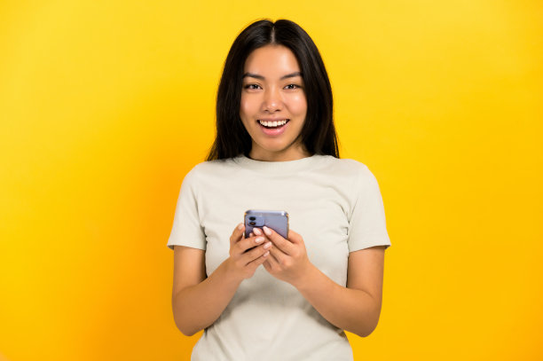 兴奋、快乐、惊讶的亚洲深色长发女孩，穿着休闲t恤，手里拿着智能手机，站在一个孤立的橙色背景上，惊讶地看着镜头图片下载