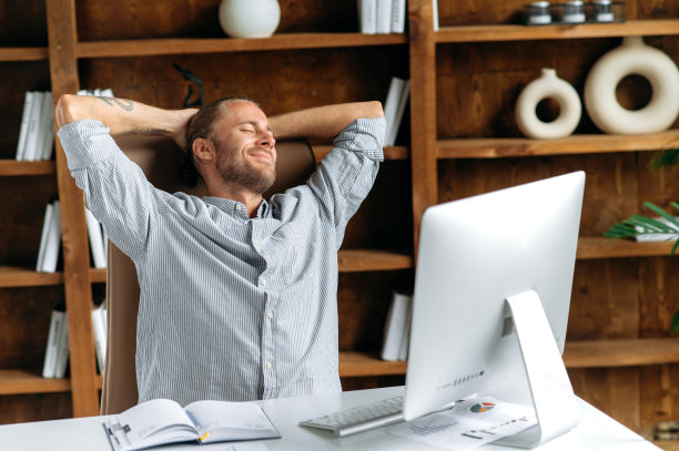 平静的年轻白人经理或自由职业者，坐在办公室的一张桌子旁，休息，把双手放在脑后，闭上眼睛，微笑着，令人不寒而栗图片下载