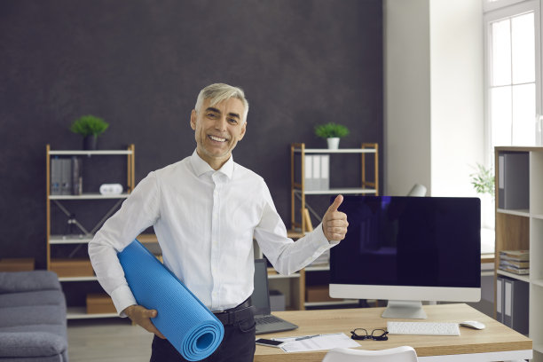 快乐健康的年长男子站在办公室，拿着瑜伽垫，给大拇指和微笑图片下载