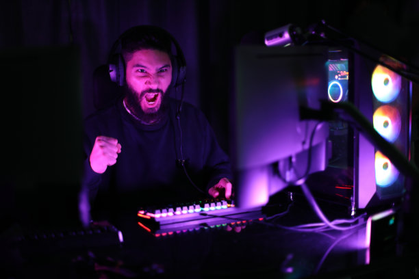 一个年轻人在一个被五颜六色的霓虹灯照亮的房间里玩电脑游戏。游戏玩法有趣且令人满意。一名男子戴着耳机，打算在互联网上玩游戏。图片下载