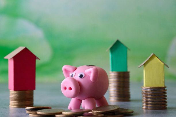 特写图像的彩色纸板房子站在成堆的硬币除了存钱罐和硬币堆，重点是前景，斑驳的绿色背景，拷贝空间，家庭财务，房地产，假日储蓄的概念图片下载