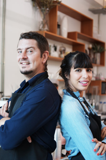 微笑的白人年轻的咖啡师夫妇的爱情或合作伙伴穿着围裙，高兴地在咖啡店工作。创业为小咖啡馆业主的理念。图片下载