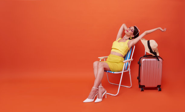 美丽的女人放松坐在沙滩椅上与手提箱在工作室夏季橙色的背景与拷贝空间。图片下载