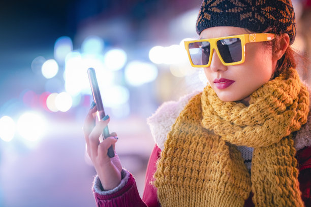 一名女子在夜晚的城市街道上用她的智能手机自拍。图片下载