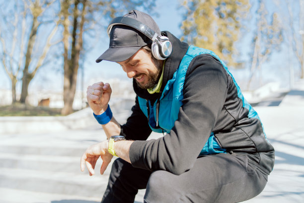 一名男运动员在晨跑时对自己的成绩很满意，微笑着，在智能手表上检查参数，高兴地握紧拳头，耳机里放着激励人心的音乐图片下载