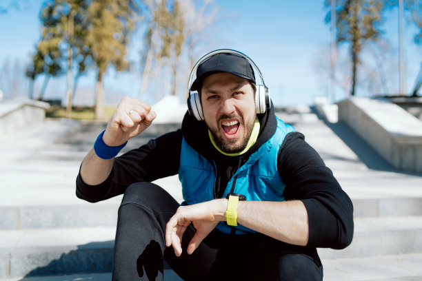 快乐的男人正坐在公园的台阶上，喜悦地喊着，拳头胜利喜悦地赢了，戴着无线耳机的男子运动员跑步者看到手表上的记录打破了公里跑图片下载