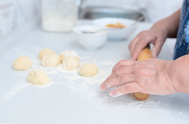 老妇人的手在白色的厨房桌子上用擀面杖擀面团，背景是模糊的磨碎的苹果和糖。有选择性的重点。制作苹果馅派的过程图片下载