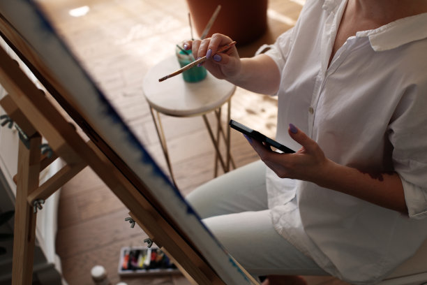 女艺术家在画架上用画笔画画，手里拿着智能手机，观看在线艺术绘画课程图片下载