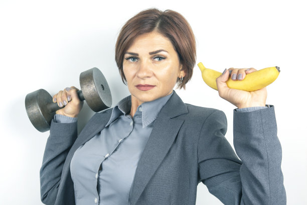 快乐美丽的商业女性在西装提起一个哑铃和一个香蕉在她的手图片下载