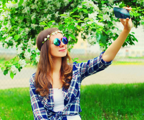 快乐微笑的年轻女性嬉皮的肖像通过智能手机戴着花的头带，太阳镜在花园或公园自拍图片下载
