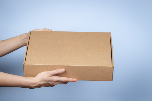 女人手里拿着棕色的天然瓦楞纸板生态包装盒。蓝色背景的邮寄包裹盒模型。包装、购物、配送理念图片下载