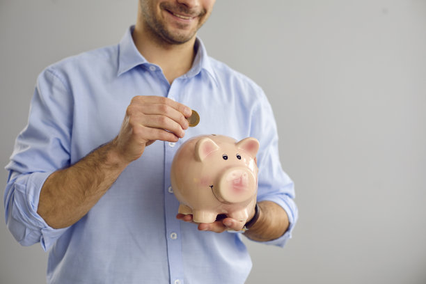 一个不知名的人站在灰色的背景上，把一枚硬币放在一个小猪形状的存钱罐里。图片下载