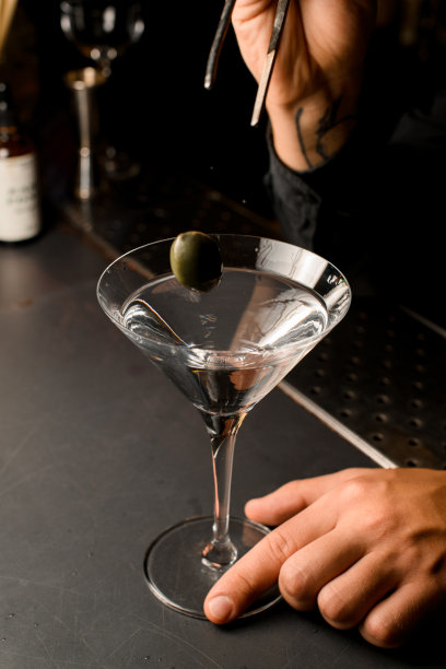 有纹身的手拿着镊子，把一个绿色的橄榄放入马提尼杯图片下载