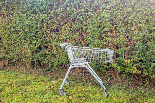 废弃的购物车靠在绿树成荫的篱笆上图片下载