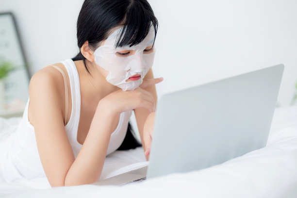 美丽年轻的亚洲女性与床单面膜躺在笔记本电脑上工作在床上的卧室，美容女孩应用面膜的皮肤皱纹在家里，皮肤护理，健康和健康。图片下载