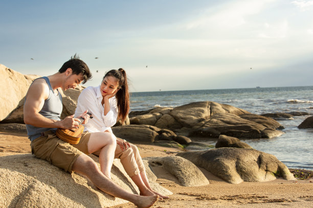 甜蜜的情侣在海边沙滩的岩石上享受尤克里里琴-图片图片下载