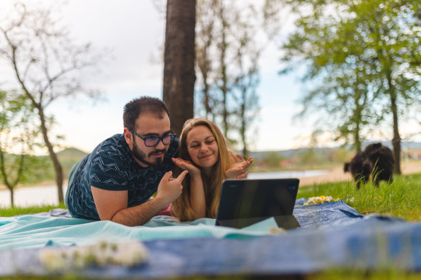 一对年轻夫妇躺在野外的野餐毯子上，同时用平板电脑进行视频通话图片下载
