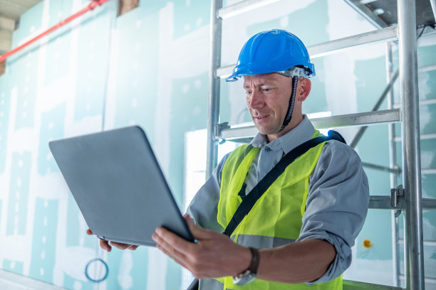 建筑师带着笔记本电脑和头盔，倚靠在建筑工地的移动脚手架上。图片下载