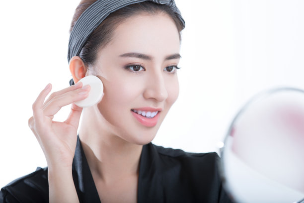 年轻的美女在化妆镜前正在上粉底，在白色的背景上-底色照片图片下载