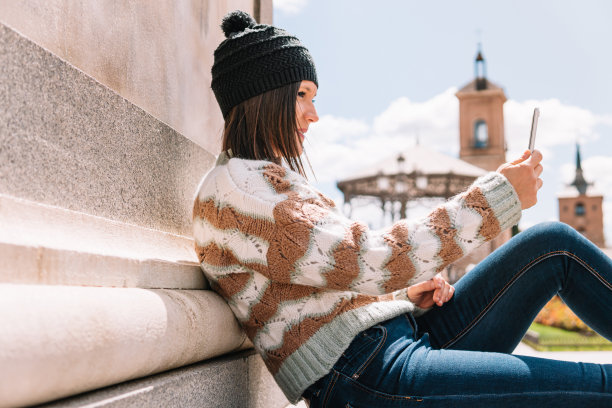 身穿棕色羊毛毛衣、头戴黑色帽子的女子坐在一座纪念碑上，一边看着她的手机。图片下载