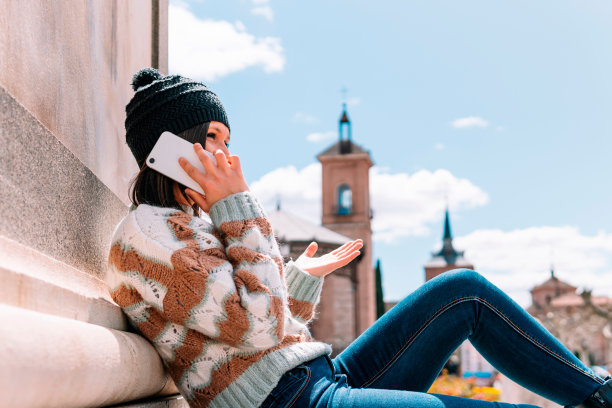 身穿棕色羊毛毛衣、头戴黑色帽子的女子坐在一座纪念碑上，一边讲手机。图片下载