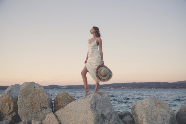 沙滩上身穿白色长裙、头戴草帽的年轻女子。时尚的暑假时尚。图片下载