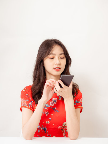 微笑的年轻亚洲女子在传统旗袍旗袍礼服使用智能手机孤立在白色背景。图片下载