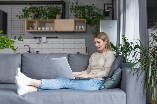 美丽的女人坐在家里的沙发上微笑着，在笔记本电脑上工作图片下载