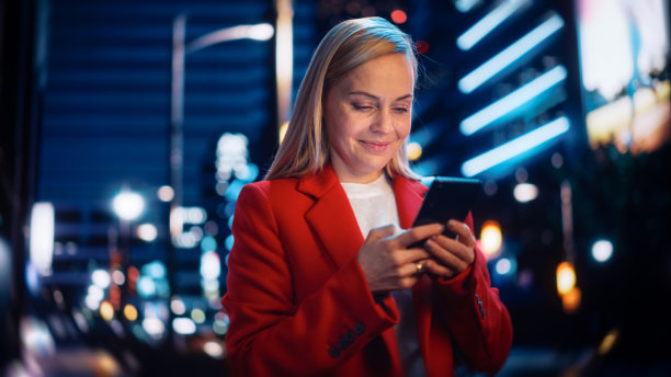 一个美丽的女人在红色大衣走在现代城市街道霓虹灯在晚上的肖像。有吸引力的女性使用智能手机和环顾城市电影环境。图片下载