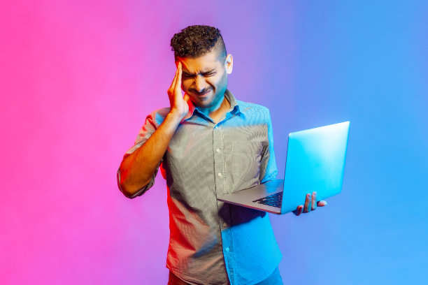 男人手持笔记本电脑，痛苦地捂着头，因过度工作和压力大的工作而患偏头痛。图片下载