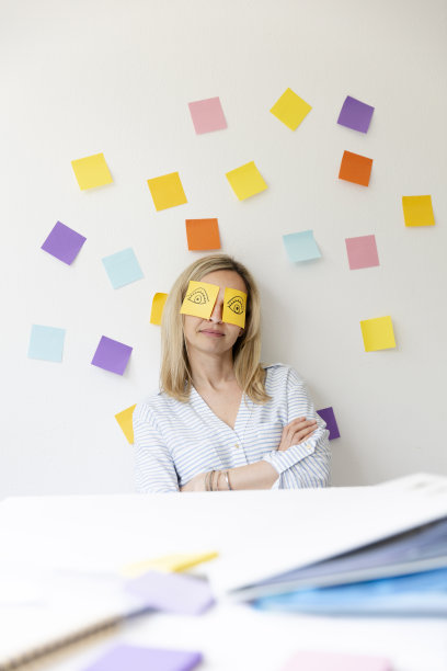 商务女性坐在明亮的现代办公室后面，她的办公桌上有许多办公室的东西，她的脸上有画着眼睛的便签和睡觉图片下载