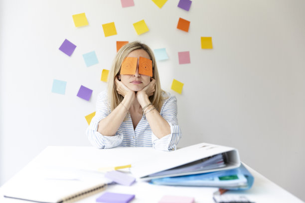 商务女性坐在明亮的现代办公室后面，她的办公桌上有许多办公室的东西，她的脸上有画着眼睛的便签和睡觉图片下载