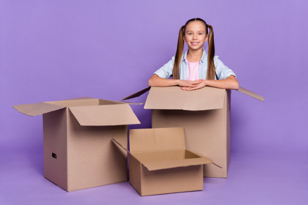 照片漂亮闪亮的小女生穿着牛仔裤衬衫坐在巨大的纸箱孤立的紫色背景图片下载