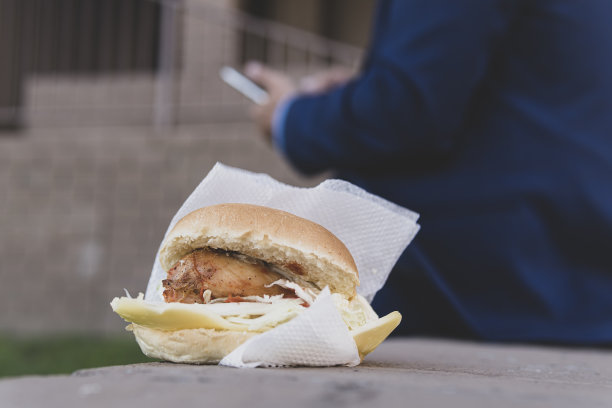鸡肉三明治躺在街上的长凳上。午餐时间吃垃圾食品的概念。商人在办公楼背景下的街道上吃着不健康的高热量食物图片下载