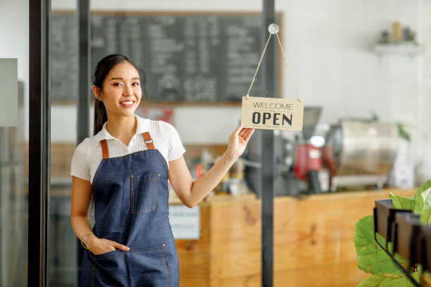身穿围裙的亚洲年轻女咖啡师拿着平板电脑站在开着招牌的咖啡馆门前。图片下载
