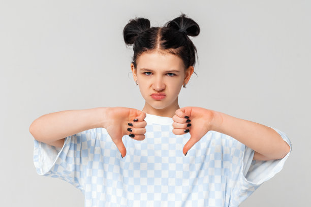 可爱的十几岁女孩看起来不开心和生气显示拒绝和消极的拇指向下的手势，站在浅灰色的背景下的时尚t恤。人们真诚的情感生活理念图片下载