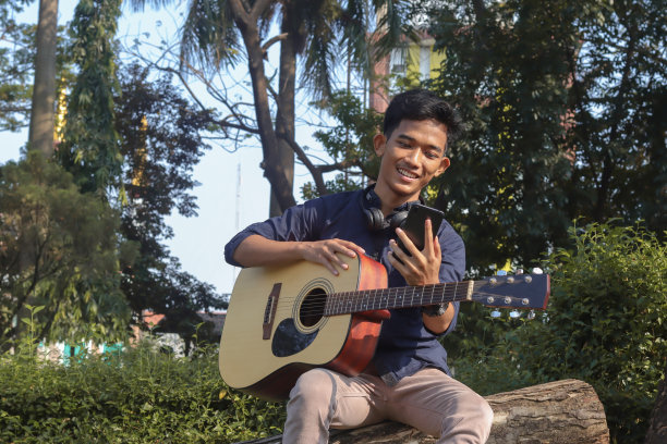 一名亚洲男子在公园玩吉他时使用手机图片下载