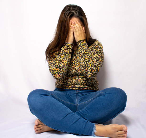 女人盘腿坐在白色背景上，双手放在脸上。提到抑郁、心理健康、成人疲劳和疲劳。图片下载