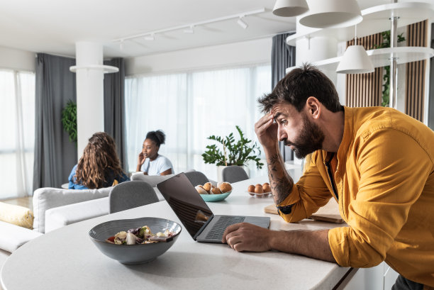 一名紧张兴奋的男子在厨房用笔记本电脑观看足球比赛现场直播，在网上下注，等待获胜结果，而他的妻子和朋友坐在客厅里。图片下载